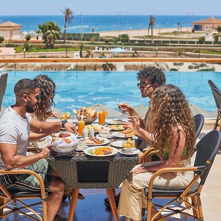 Tolip Resort El Galala Majestic Ain Sukhna Eksteriør bilde The photo shows a group of five people enjoying a meal together outdoors, likely at a restaurant or cafe. They are seated at a table with a beautiful view of a swimming pool and the sea in the background. The table is filled with various dishes, and 