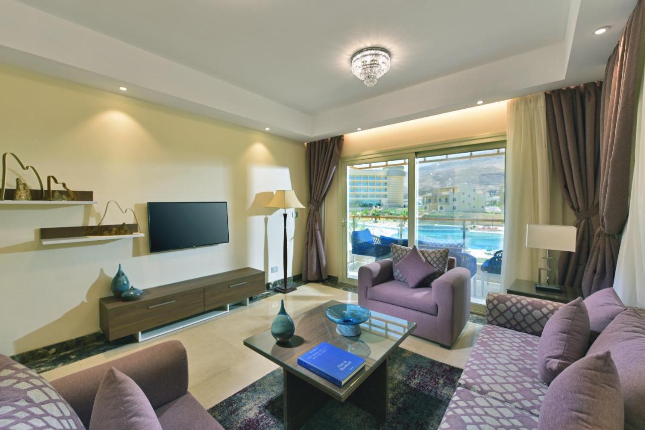 Tolip Resort El Galala Majestic Ain Sukhna Eksteriør bilde The photo shows a modern living room that features a stylish and cozy design. There are two purple sofas with decorative cushions arranged around a coffee table. The coffee table has a blue book and a decorative item on it. A flat-screen TV is mounte