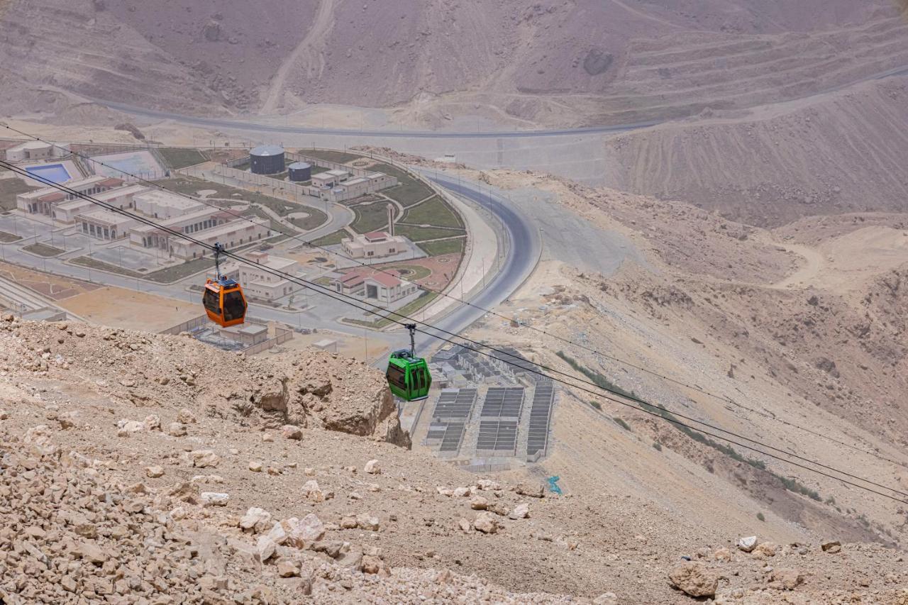 Tolip Resort El Galala Majestic Ain Sukhna Eksteriør bilde The photo shows a scenic view from a high elevation, featuring a cable car system. Two cable cars are visible, one orange and one green, traveling along cables down the mountainside. Below, there appears to be a developed area with roads and light st