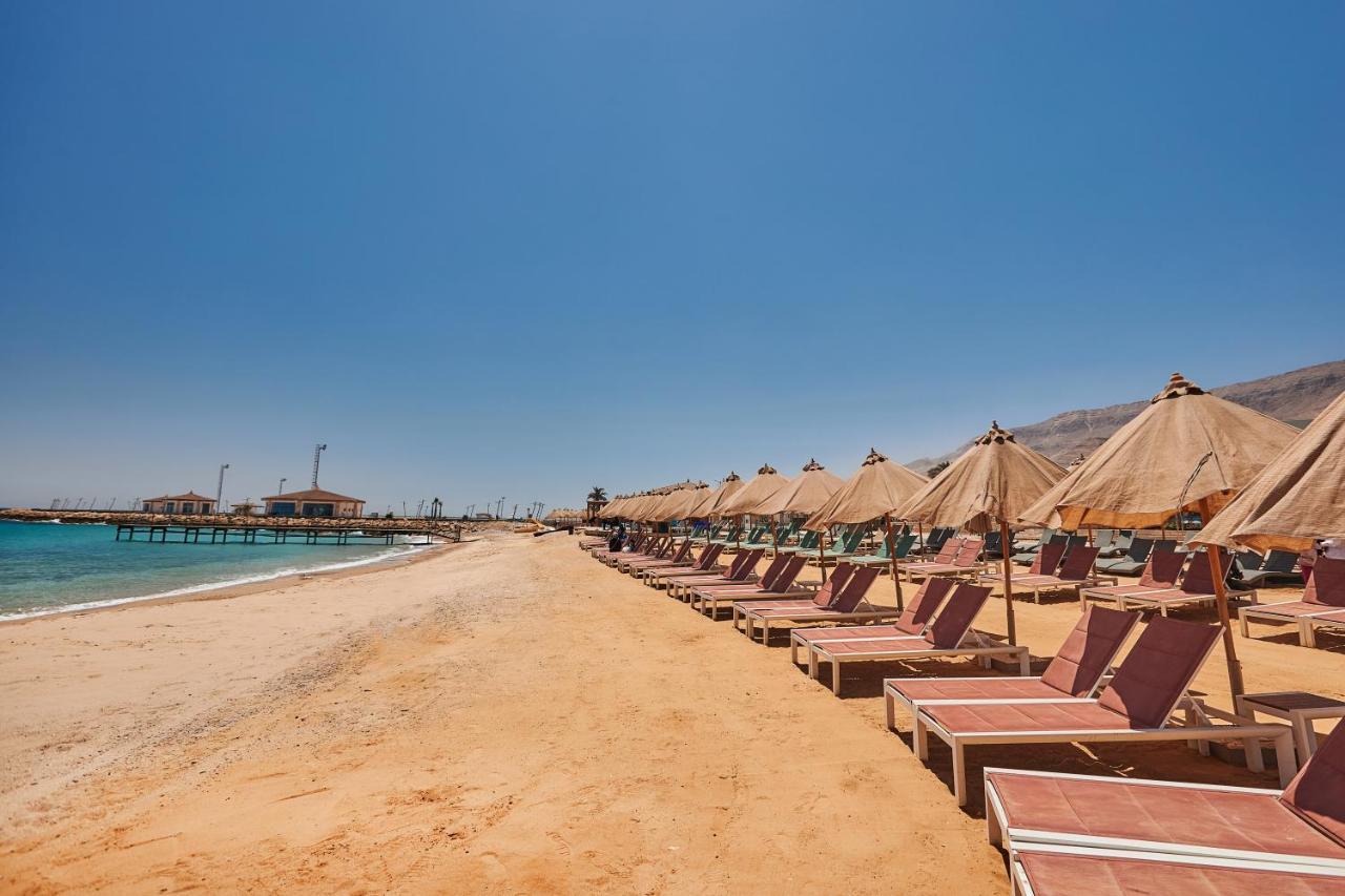 Tolip Resort El Galala Majestic Ain Sukhna Eksteriør bilde The photo shows a scenic beach with a stretch of golden sand under a clear blue sky. There are several beach loungers arranged in rows, each accompanied by thatched umbrellas providing shade. In the background, there is a wooden pier extending into t