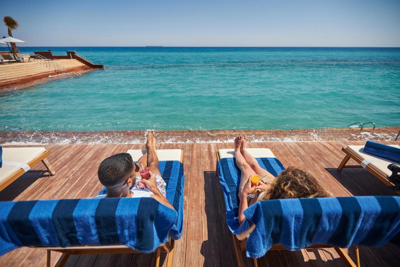 Tolip Resort El Galala Majestic Ain Sukhna Eksteriør bilde The photo depicts two people relaxing on lounge chairs by the waterfront. They are positioned on a wooden deck with a view of the clear, turquoise water. Both individuals are reclining, enjoying the sun, and each has a drink in hand. The scene convey