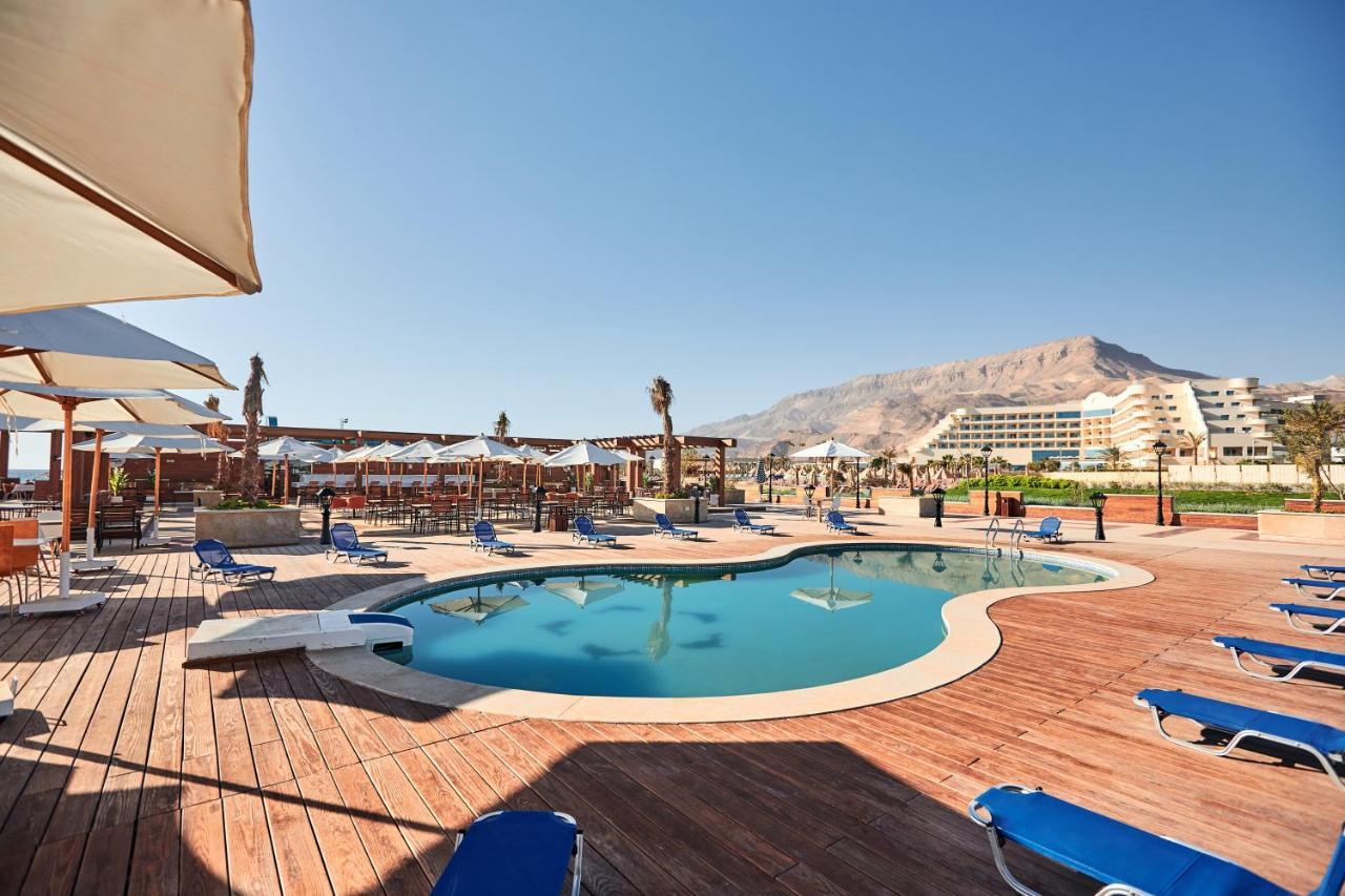 Tolip Resort El Galala Majestic Ain Sukhna Eksteriør bilde The photo shows a swimming pool area surrounded by sun loungers and umbrellas. The pool has a unique, curvy shape and is situated on a wooden deck. In the background, there are several buildings, likely part of a resort or hotel, and a mountainous la