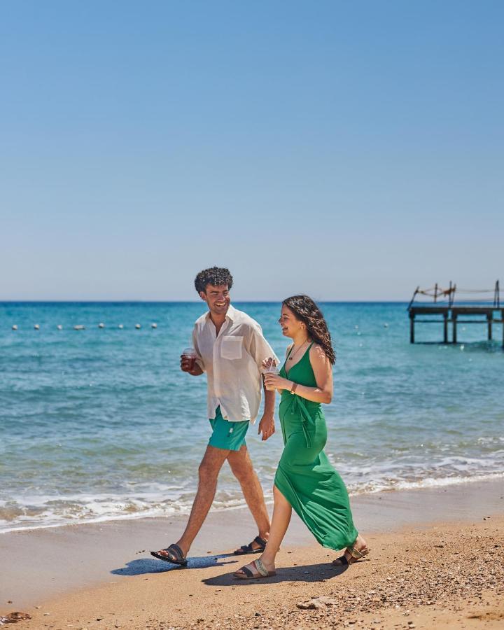 Tolip Resort El Galala Majestic Ain Sukhna Eksteriør bilde The photo shows a couple walking along a beach. The man is wearing a white short-sleeved shirt and green shorts, while the woman is dressed in a flowing green dress. They both appear to be enjoying the pleasant weather, with the ocean in the backgrou