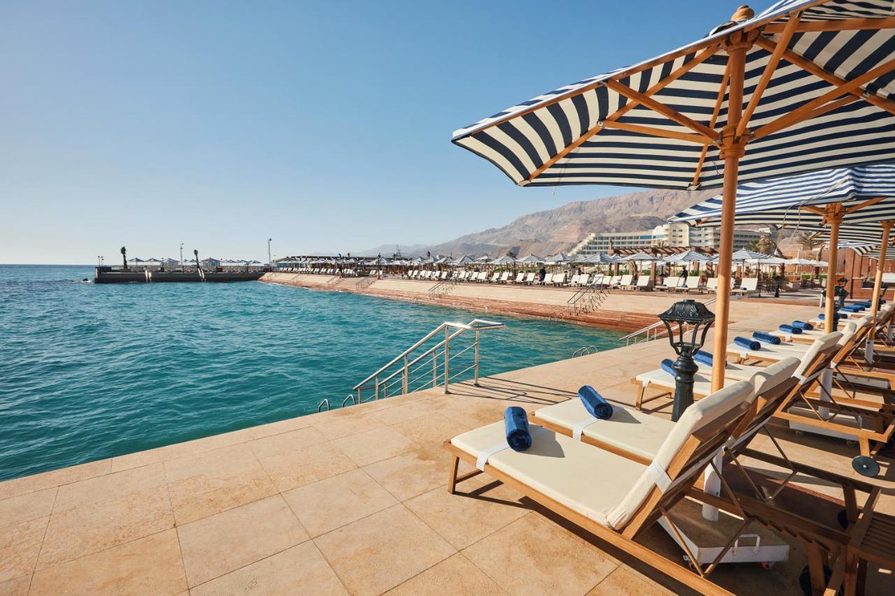 Tolip Resort El Galala Majestic Ain Sukhna Eksteriør bilde The photo shows a scenic beachside area with clear blue water. In the foreground, there are several sun loungers set up on a tiled surface, with blue towels neatly rolled on each one. The loungers are shaded by striped umbrellas. In the distance, a w