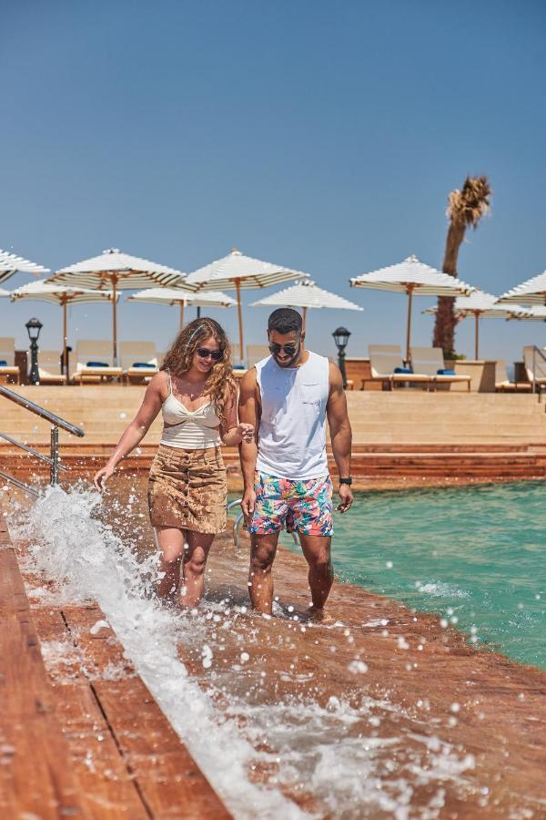 Tolip Resort El Galala Majestic Ain Sukhna Eksteriør bilde The photo depicts a couple walking along a poolside area. They are enjoying a sunny day near a swimming pool, with splashes of water around them. The woman is wearing a light-colored top and a patterned skirt, while the man is dressed in a sleeveless