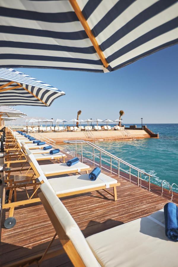 Tolip Resort El Galala Majestic Ain Sukhna Eksteriør bilde The photo depicts a serene outdoor area by the waterfront. There are several lounge chairs arranged in a row, each equipped with blue towels. Large, striped umbrellas provide shade over some of the chairs. In the background, there is a wooden deck th