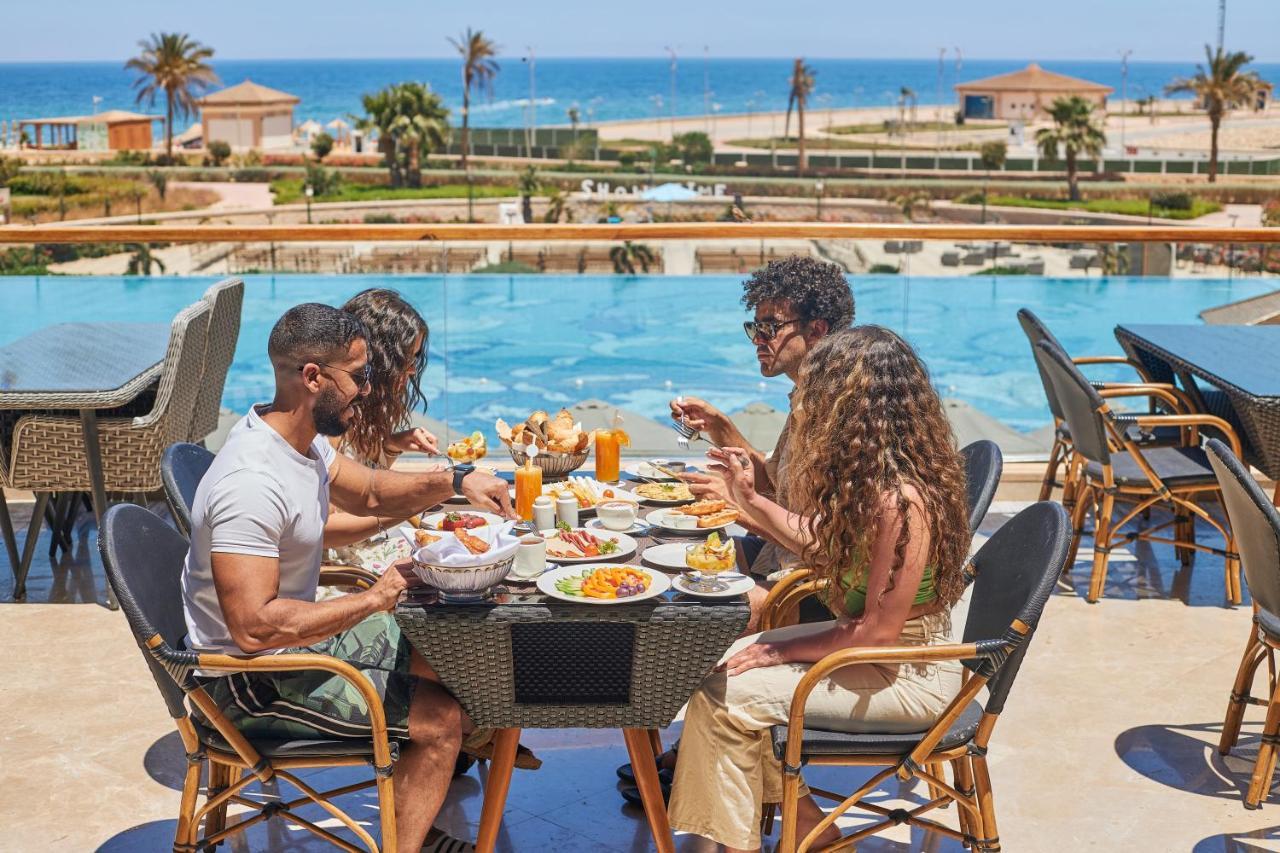 Tolip Resort El Galala Majestic Ain Sukhna Eksteriør bilde The photo shows a group of five people enjoying a meal together outdoors, likely at a restaurant or cafe. They are seated at a table with a beautiful view of a swimming pool and the sea in the background. The table is filled with various dishes, and 