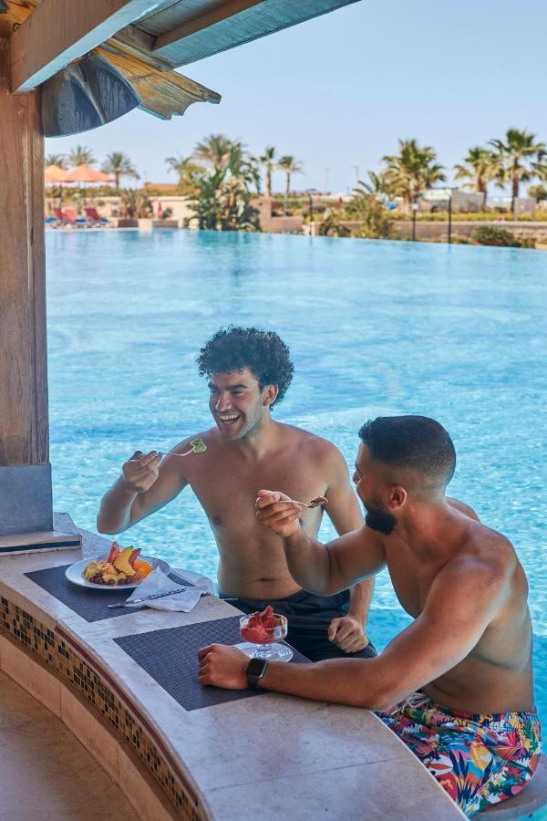 Tolip Resort El Galala Majestic Ain Sukhna Eksteriør bilde The photo features two young men enjoying a meal together. They are sitting at a poolside table with a clear blue swimming pool in the background. The men are shirtless, suggesting a casual, relaxed atmosphere, likely in a warm environment. One man i