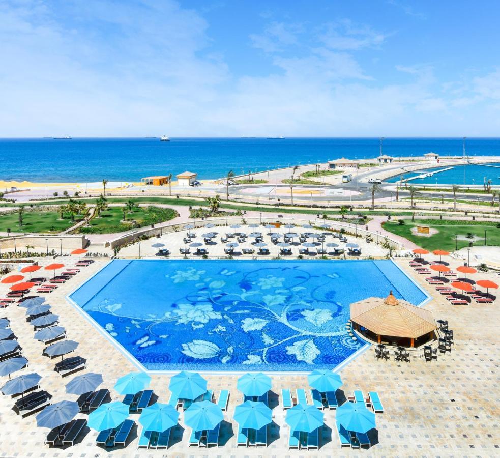 Tolip Resort El Galala Majestic Ain Sukhna Eksteriør bilde The photo features a large swimming pool with a decorative design on its surface, surrounded by sun loungers and umbrellas. In the background, there is a view of the sea, with a clear blue sky and a few scattered clouds. The area is well-maintained, 