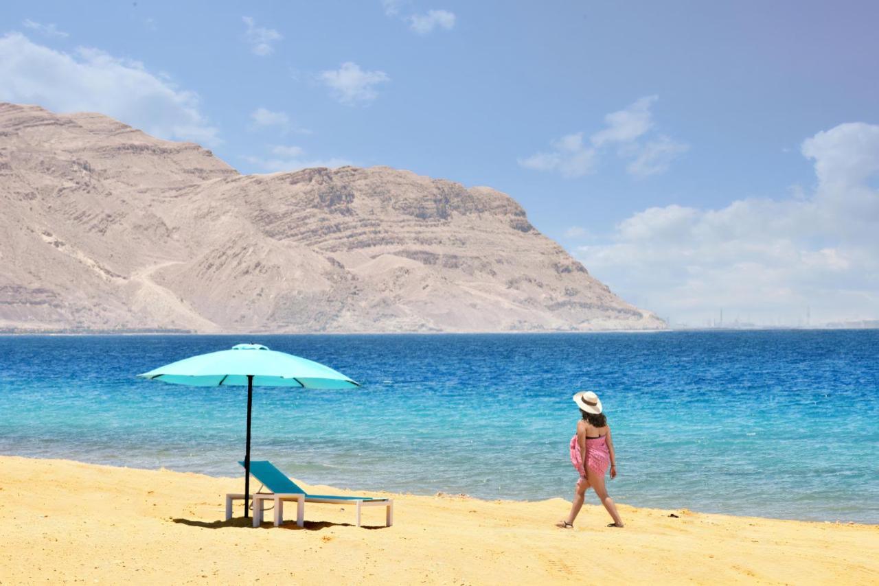 Tolip Resort El Galala Majestic Ain Sukhna Eksteriør bilde The photo depicts a serene beach scene. In the foreground, there is a sandy shore with a single beach chair and an umbrella in a light blue color. A person dressed in a pink outfit is walking along the shoreline, enjoying the beach atmosphere. In the